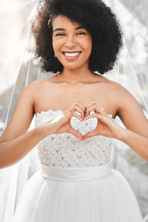 Were celebrating true love today. Portrait of a happy and beautiful young bride making a heart shape with her hands outdoors on her wedding day.の写真素材