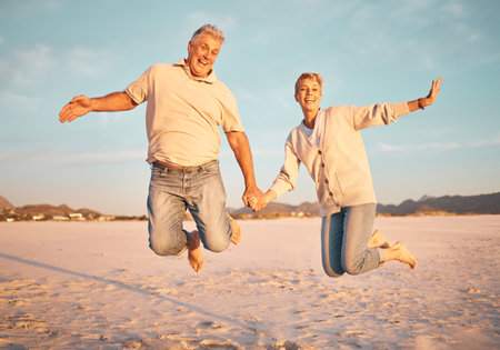 Couple, beach and active seniors hold hands while jumping in sand, happy and excited at sunset. Love, family and freedom with mature man and woman jump in celebration of retirement, travel and energyの写真素材