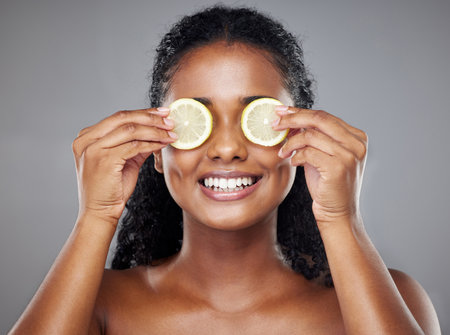 Skincare, portrait and woman with lemon on her eyes for health, beauty and wellness standing in a studio. Happy, smile and face of girl model from Brazil with citrus fruit isolated by gray backgroundの写真素材