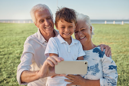 Kid, grandparents and phone selfie on grass in park spending time together on the weekend. Grandma, grandpa and boy child with smile on face, happy family take outdoor portrait on smartphone on fieldの写真素材