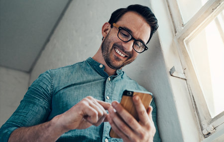Now thats the kind of text I like to see. a handsome young businessman smiling while using a smartphone in a modern office.の写真素材