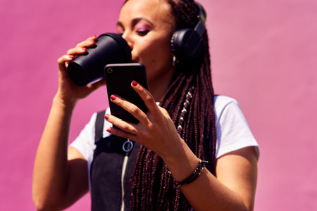Coffee and tech are millennials necessities. an attractive young woman standing against a pink wall alone and drinking coffee while using technology.の写真素材