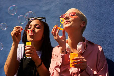 Blowing bubbles makes us happy. two attractive young women sitting against a blue wall together and bonding by blowing bubbles.の写真素材