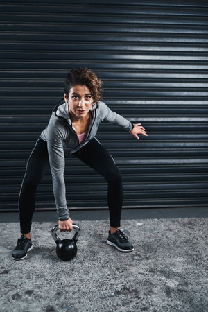When it burns, push harder. Full length shot of an attractive young woman exercising against a black background and lifting kettle bells.の写真素材