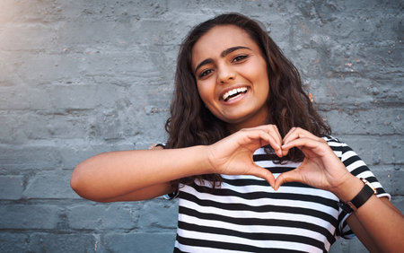 My heart is packed full of love for you. Portrait of a young woman making a heart shape with her hands against a grey wall.の写真素材