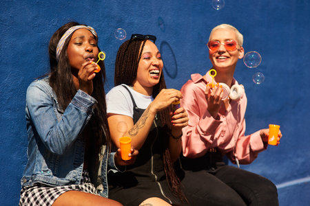 Bubbles, bubbles everywhere. three attractive young women sitting against a blue wall together and bonding by blowing bubbles.の写真素材