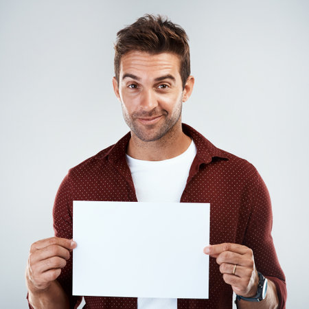 Portrait of a cheerful young man holding and displaying a poster while standing against a grey background.の写真素材