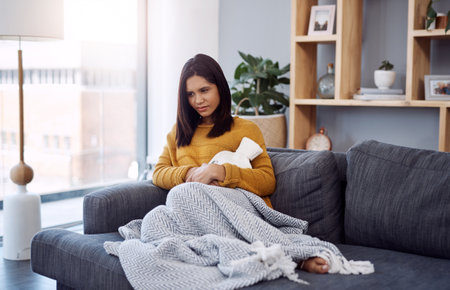 Full length shot of an attractive young woman suffering from stomach cramps while sitting on a sofa at home.の写真素材