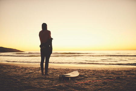 Out for some fun adventures in the sun. Rearview of a young surfer putting on a wetsuit at the beach.の写真素材