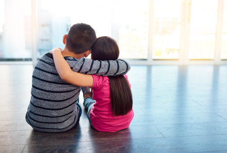 Keep the family close. Rearview shot of two young children sitting on the floor and posing with their arms around each other at home.の写真素材
