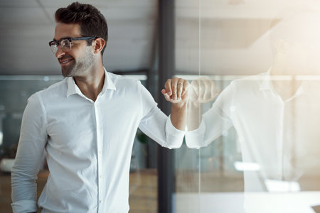 Modern man in a modern office. a handsome young businessman leaning against a glass wall in his modern office.の写真素材