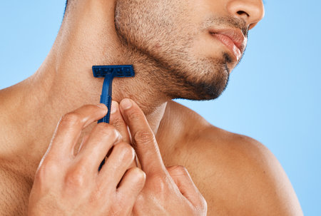 Man, hands and shaving with razor for beard grooming, skincare or facial treatment against a blue studio background. Hand of male in beauty shave for face, skin or cosmetic care for clean hygieneの写真素材