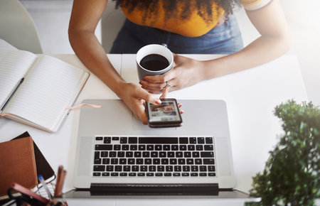 All set for the day, connected with coffee. High angle shot of an unrecognizable female designer using a cellphone in her home office.の写真素材
