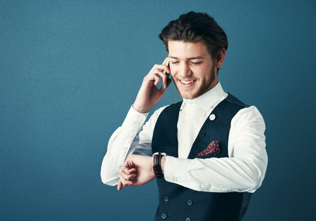 I can be there in 25 minutes. Studio shot of a handsome young businessman checking the time while making a phonecall against a blue background.の写真素材