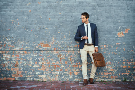 Waiting for his ride. Full length shot of a handsome young businessman standing against a grey facebrick wall.の写真素材
