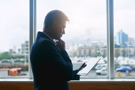 Working in a global and mobile business world. a mature businessman working on a digital tablet while standing at a window in an office.の写真素材