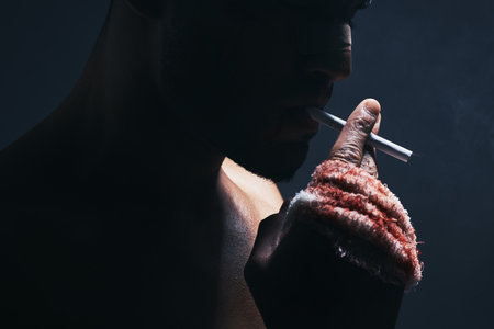 Injury, cigarette and blood bandage on hand of kickboxing man in studio for shadow, art deco and bad habits background. Male champion boxer smoking after fighting in MMA violence sports competitionの写真素材