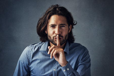 Can you keep a secret. Studio portrait of a handsome young man posing with his finger on his lips against a grey background.の写真素材