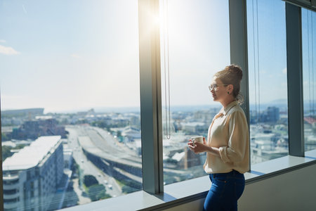 She has one great view from her office. a confident young businesswoman drinking coffee while standing next to a window and looking through it inside of the office.の写真素材