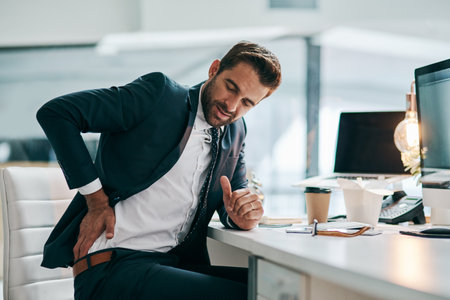 His back isnt playing along today. a uncomfortable young businessman holding his back in discomfort due to pain while being sated in the office.の写真素材