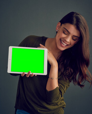 The device can present itself too. Studio shot of an cheerful young woman holding a digital tablet while standing against a dark background.の写真素材