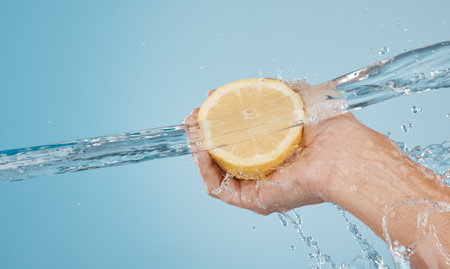 Water, splash and beauty with hands and lemon for health, wellness and hydration. Vitamin c, refreshing or nutrition with citrus fruits and model against blue background for natural, vitality or dietの写真素材