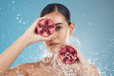 Beauty, skincare and woman with pomegranate in face, splash and natural treatment for fresh, clean and glowing skin. Water, fruit and healthy cosmetics for beautiful model girl with studio backgroundの写真素材