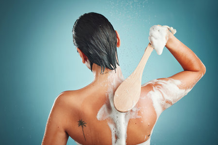 Skincare, back and woman in a shower for cleaning, hair and wellness against a blue background mockup. Beauty, brush and skin, scrub and exfoliate by girl model in a studio, relax and washingの写真素材