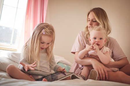 Family time in a digital world. a little girl using a digital tablet while bonding with her mother and sister on the bed.の写真素材
