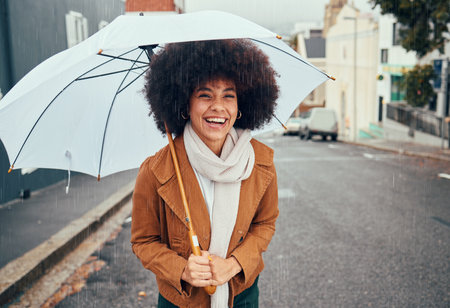 Rain, umbrella and city with a walking black woman in the street during a cold or wet winter day. Water, insurance and cover with a young afro female outdoor for a walk in an urban town roadの写真素材