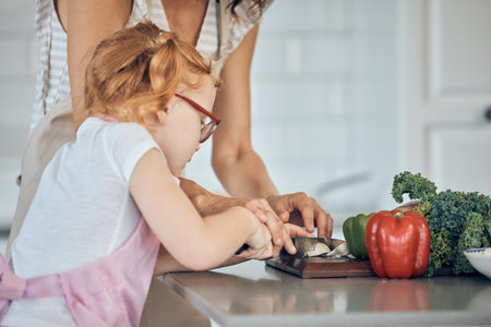 Mother teaching, child cutting vegetable and learning skill in home kitchen together on counter. Girl knife focus, cooking education with mom teacher and cut vegetables for dinner food in houseの写真素材