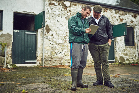 Look at this new tractor on the market. two cheerful young farmers busy on a digital tablet while...