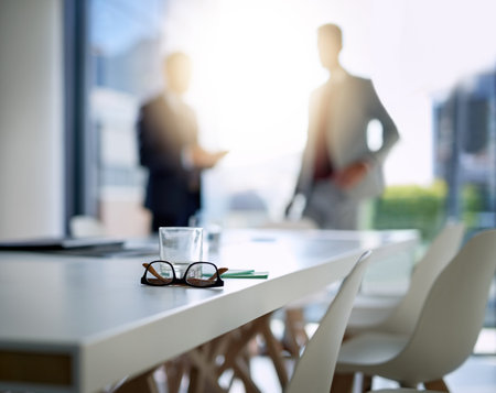 Where business excellence happens. glasses and a glass on a boardroom desk with two businessmen blurred in the background.の写真素材