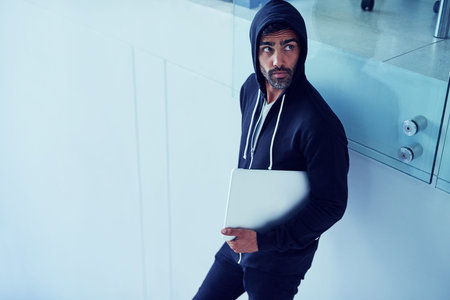 Getting up to no good. a shady-looking young man holding a laptop in an office.