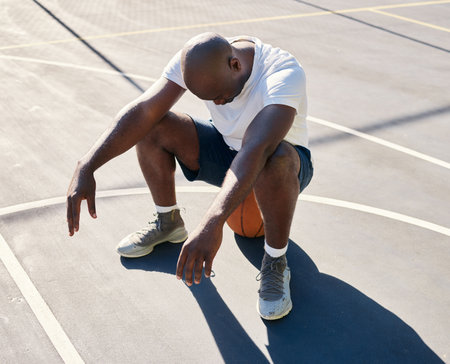 Basketball court, tired and black man sitting on a basketball resting or relaxing on a fitness training or workout break. Loser, wellness and African sports athlete with fatigue in an exercise gameの写真素材