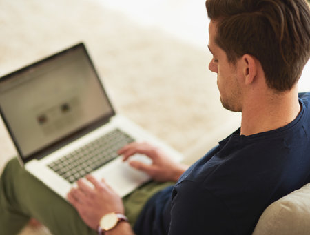 Doing a little browsing at home. High angle shot of a young man using his laptop while sitting on the sofa at home.の写真素材