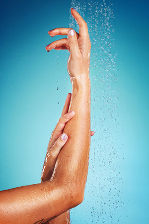 Arm, water and person in the shower cleaning body against a blue studio background. Spa, wellness and hand of model woman with luxury hydration from liquid, dermatology and healthy care for skinの写真素材