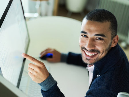Portrait of a smiling young man sitting at a desk working on a computer with dual monitors.の写真素材