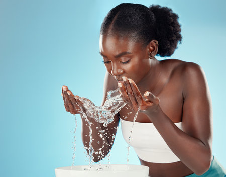 Water, wash and woman splashing her face for cleansing hygiene and skincare on a blue studio background. Skin care, body care and beauty female washing her for facial health and wellnessの写真素材
