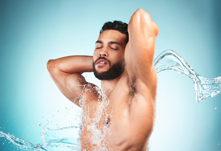 Beauty, shower and water with a man model in studio on a blue background for hygiene or hydration. Wellness, cleaning and splash with a handsome young male washing or cleansing in the bathroom aloneの写真素材