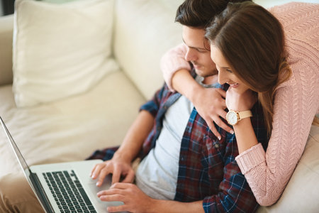 Wanna download a movie for tonight. a happy young couple using a laptop together while relaxing on the sofa at home.の写真素材