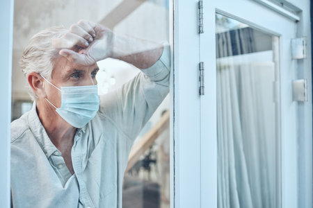Lonely, face mask and senior man in the house for quarantine, protection and isolation during pandemic. Depression, frustrated and sad elderly guy with covid looking outside the window at his home.の写真素材