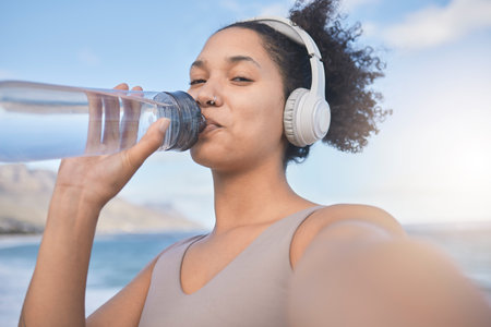 Woman runner, drinking water and selfie with music while outdoor for exercise, workout or fitness. Black woman, headphones and beach for training, wellness or health in nature by ocean in summer sunの写真素材