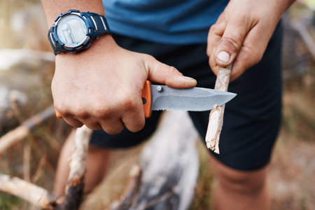 Hands, knife and cutting with a hiking man carving a stick outdoor in nature while camping for adventure. Wood, weapon and tool with a male camper slicing a twig in the mountains for survivalの写真素材