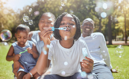 Happy, nature and black family blowing bubbles while playing, bonding and enjoying summer in the park. Happiness, father and mother with children having fun together in a green garden in South Africaの写真素材