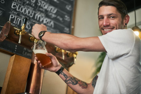 Fancy a cold one. Portrait of a young bartender pouring beer from a tap behind the bar.