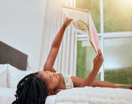 Kid reading story book in bed, relax and enjoy fairytale for entertainment, child development and literature. Story time, youth life growth and learning black girl read cartoon books in bedroomの写真素材