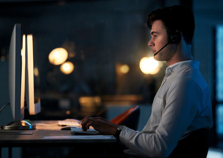 Call center, night office and employee typing, working late shift or overtime for commission. Crm, contact us and telemarketing, sales agent or support worker in dark office consulting on computer.の写真素材