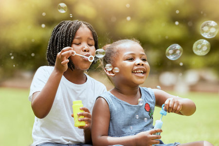Black kids, children and blowing bubbles at park, having fun and bonding. Girls, happy sisters and playing with soap bubble toys, relax and enjoying garden together outdoors in nature on grassの写真素材