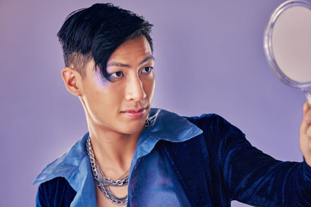 Asian man with glitter makeup, mirror for creative beauty and lgbt gender performance in fantasy art. Purple background in studio, punk fashion jewellery in Seoul and proud of unique fluid identityの写真素材